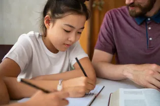 Young student with her brunette hair tied up. She is writing with a pencil in her notebook and is sitting next to a coach. He is mostly out of the picture, but he has a beard, and is wearing a burgundy top with a blue collar.